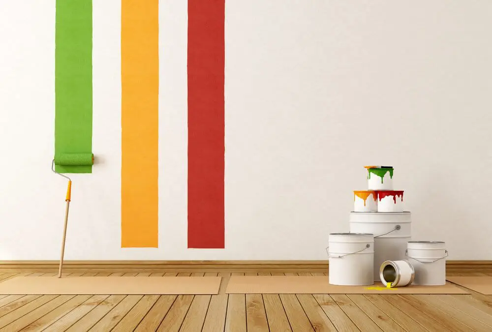 Bright green, yellow, and red stripes on a white wall, with a paint roller leaning on cardboard. Paint buckets sit on the wooden floor, one spilling over.