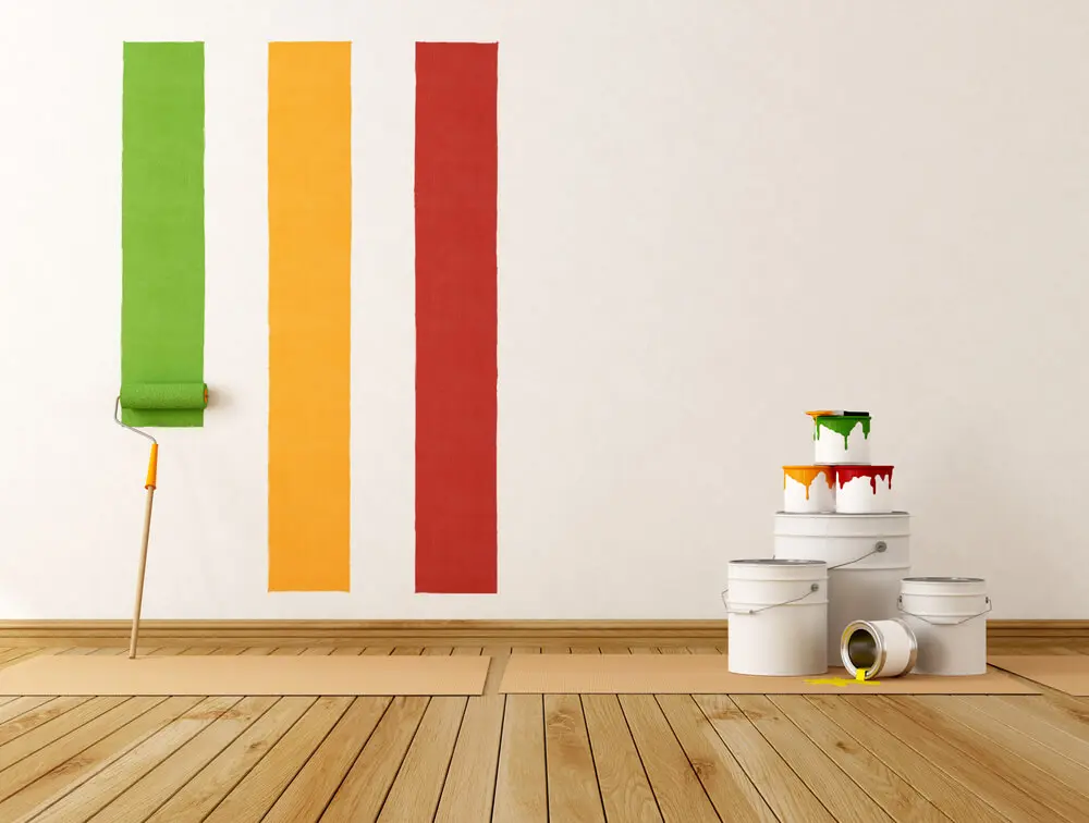 Bright green, yellow, and red stripes on a white wall, with a paint roller leaning on cardboard. Paint buckets sit on the wooden floor, one spilling over.