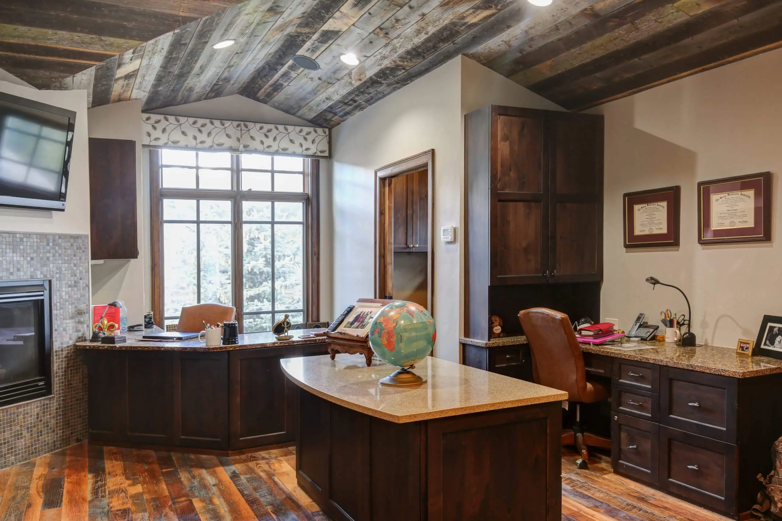 40 A home office with a globe, multiple desks, a large window, high wooden ceilings, a wooden floor and wooden cabinets.