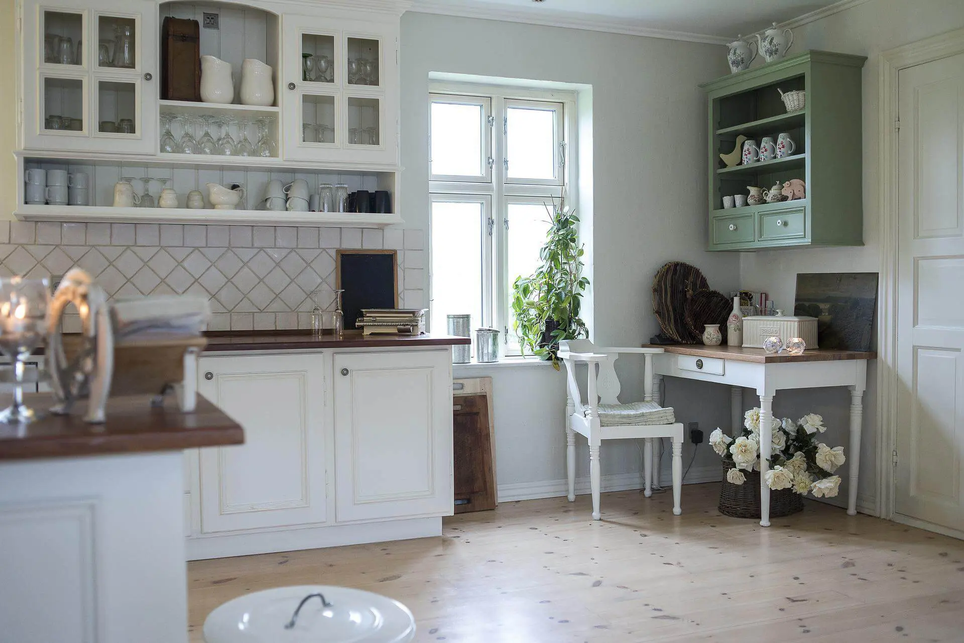 bookcase-3532950_1920 (1) An older style kitchen with white walls and white and green cabinets, with older, rustic style furniture and a wooden floor.