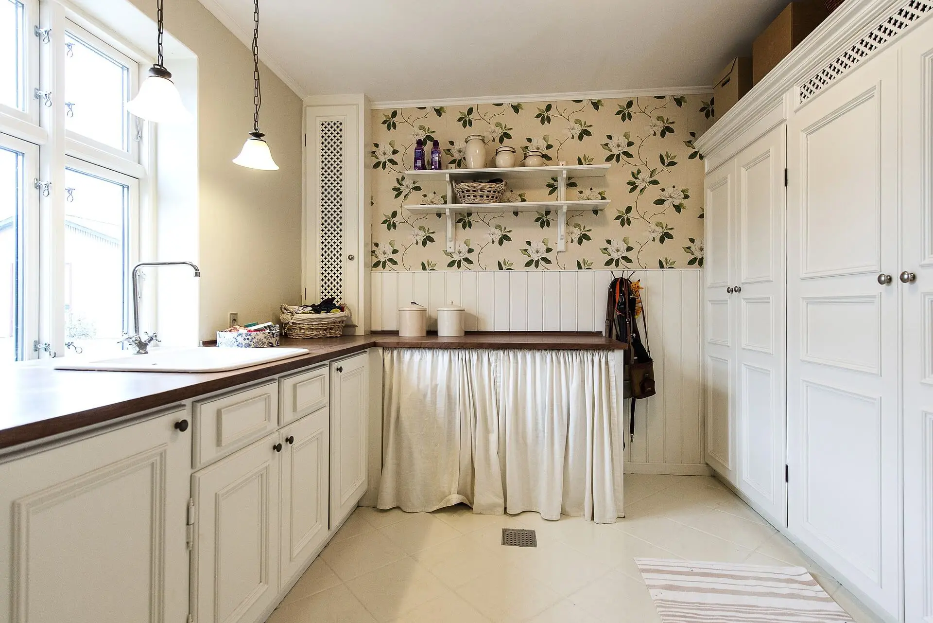 A perspective view of a kitchen in all white with dark wood countertops and flowery wallpaper.