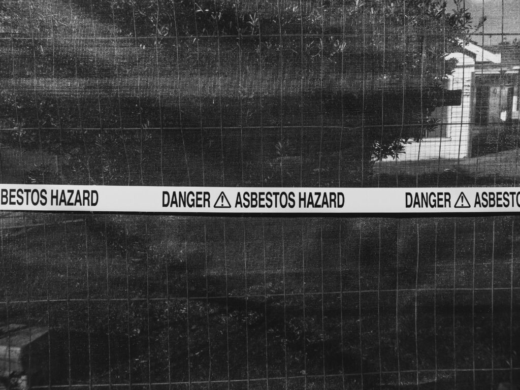 Asbestos warning tape around the fence of a home in a black and white photograph with a tree blocking most of the view.