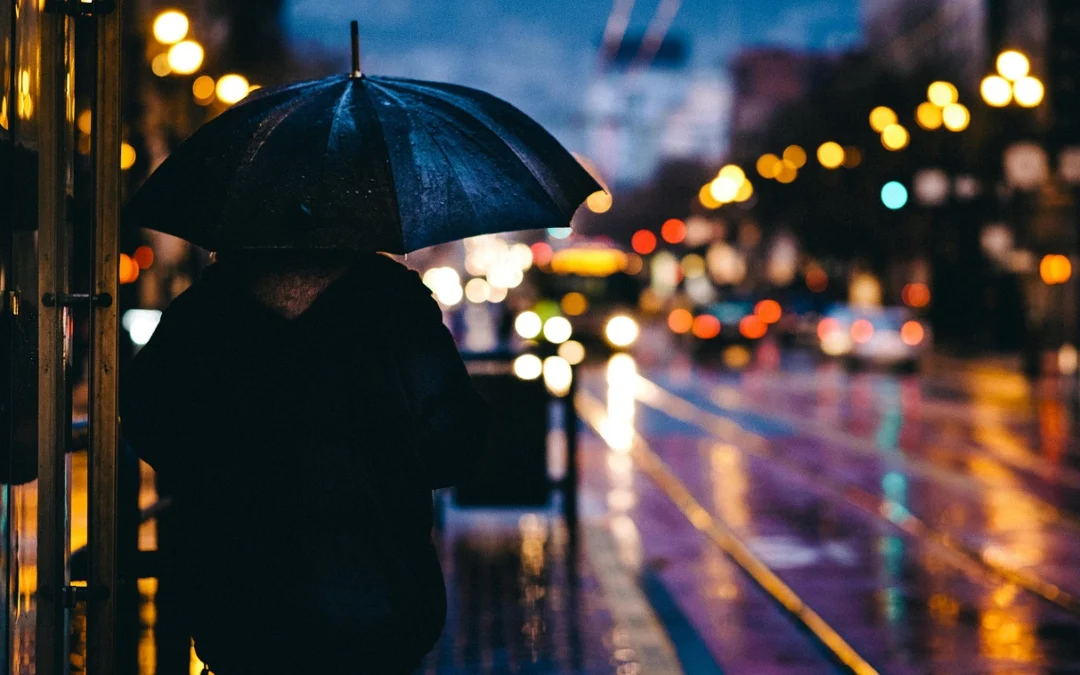 to the left a person is holding an umbrella, looking away from the camera. In the distance is unfocused street lights and car lights and a stretch of wet pavement reflecting them.