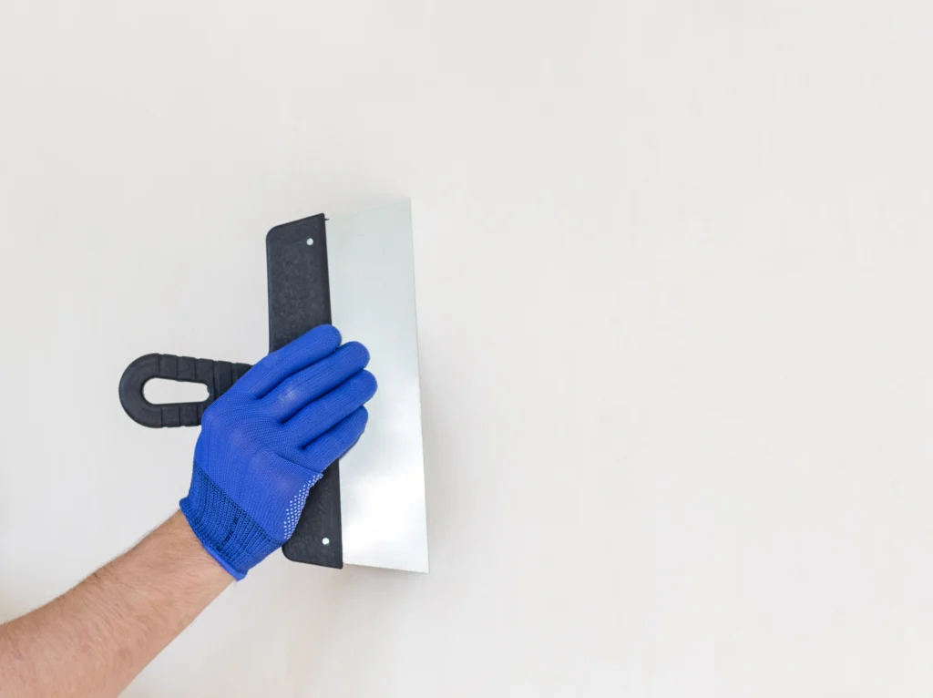 A hand holding a trowel to skim coat a surface and prepare it for painting on a white wall.